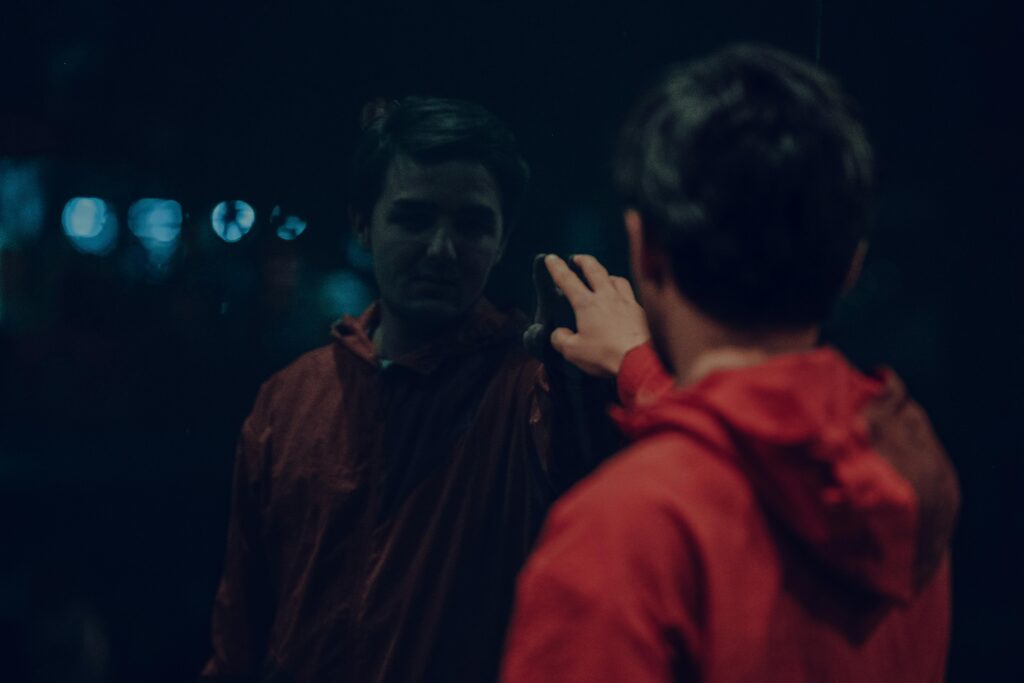 pexels-photo-11067005-11067005 A man in a red jacket looks at his reflection in a dark mirror at night.
